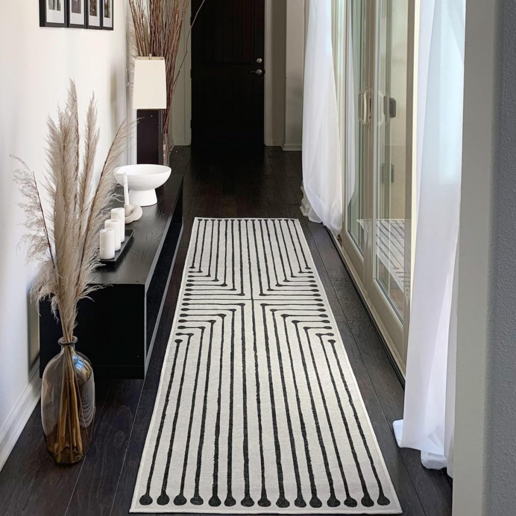 A narrow hallway with a dark floor boasts an Amazon Prime delivery on a black and white geometric rug, alongside a sleek black console table adorned with candles and decor, all beautifully complemented by sheer curtains on the right.