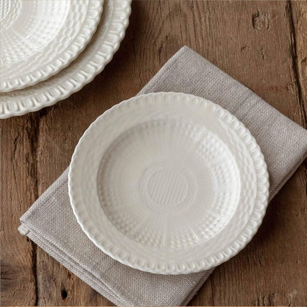 A white ceramic plate with a textured pattern rests on a folded gray napkin atop a wooden surface, inspiring elegant Friendsgiving ideas. Additional plates are partially visible in the background.