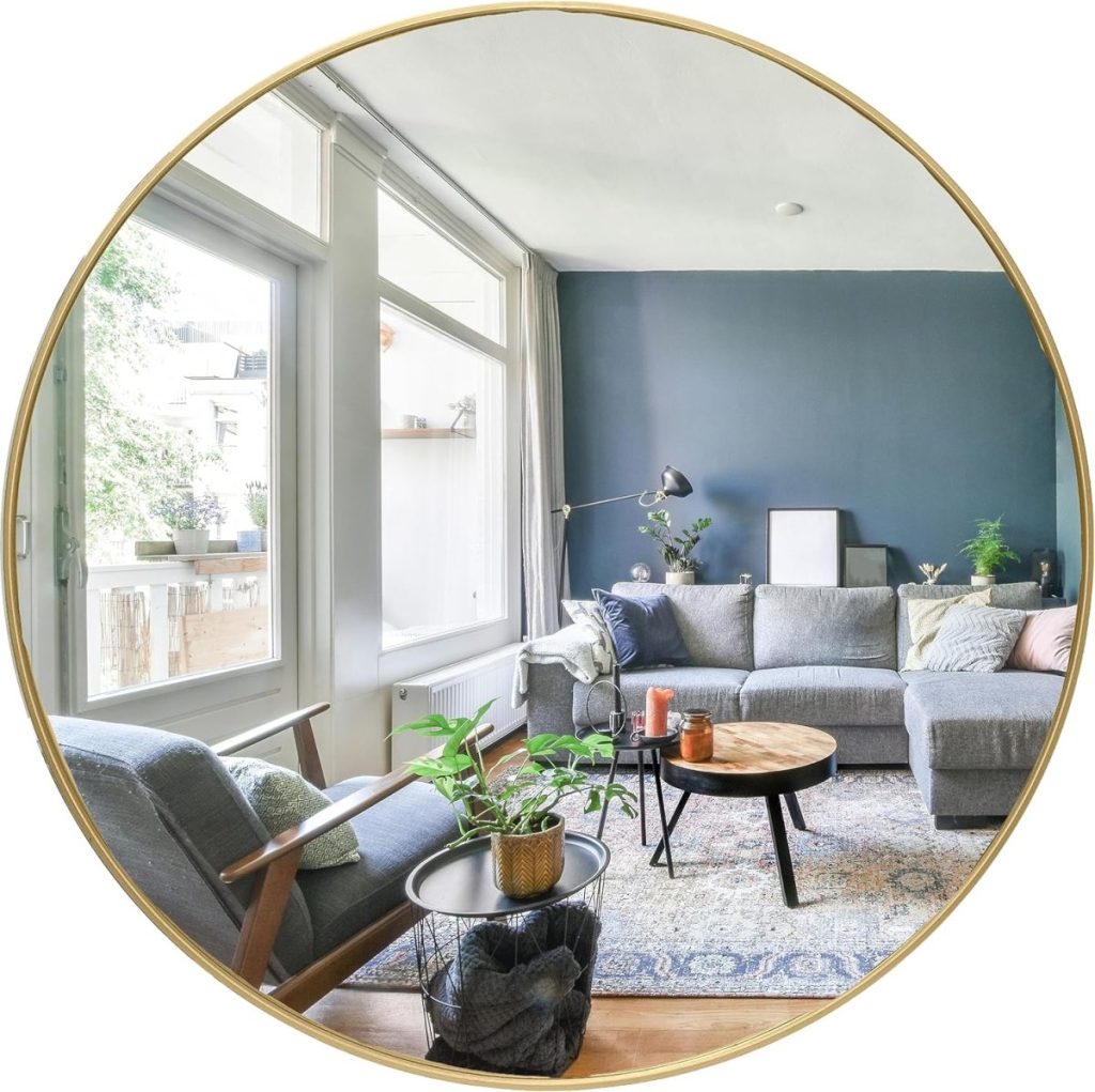 A cozy living room with a sectional sofa, armchair, round coffee table, and indoor plants. The room features large windows with natural light and a blue accent wall.