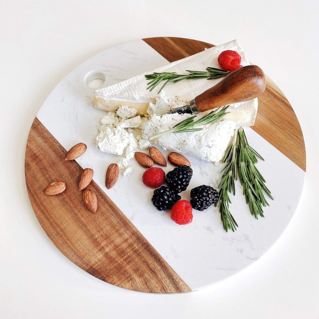 A round cutting board features baked brie with jam, a cheese knife, almonds, blackberries, raspberries, and rosemary sprigs.