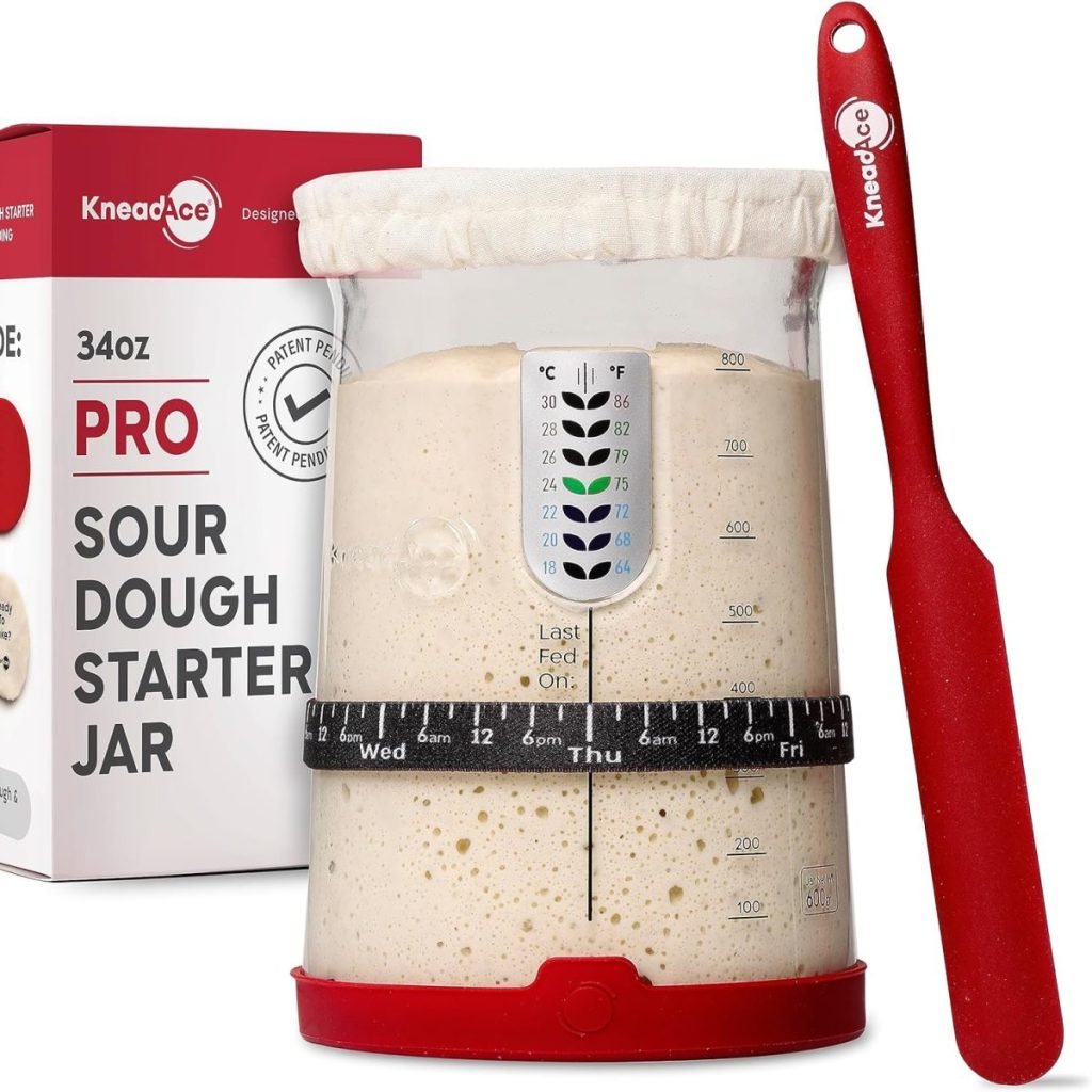 Sourdough starter jar with measurement markings, a cloth cover, and a red spatula. Box labeled "KneadAce Pro" in the background.