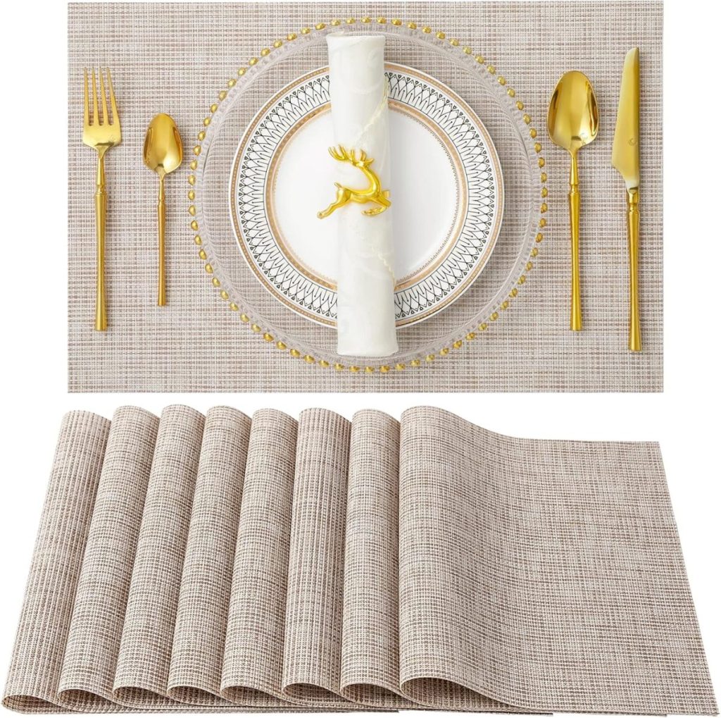 Elegant table setting with gold cutlery, a decorative plate, and a rolled napkin on a beige woven placemat. A stack of similar placemats is displayed below, accompanied by sleek glass water bottles for an added touch of sophistication.