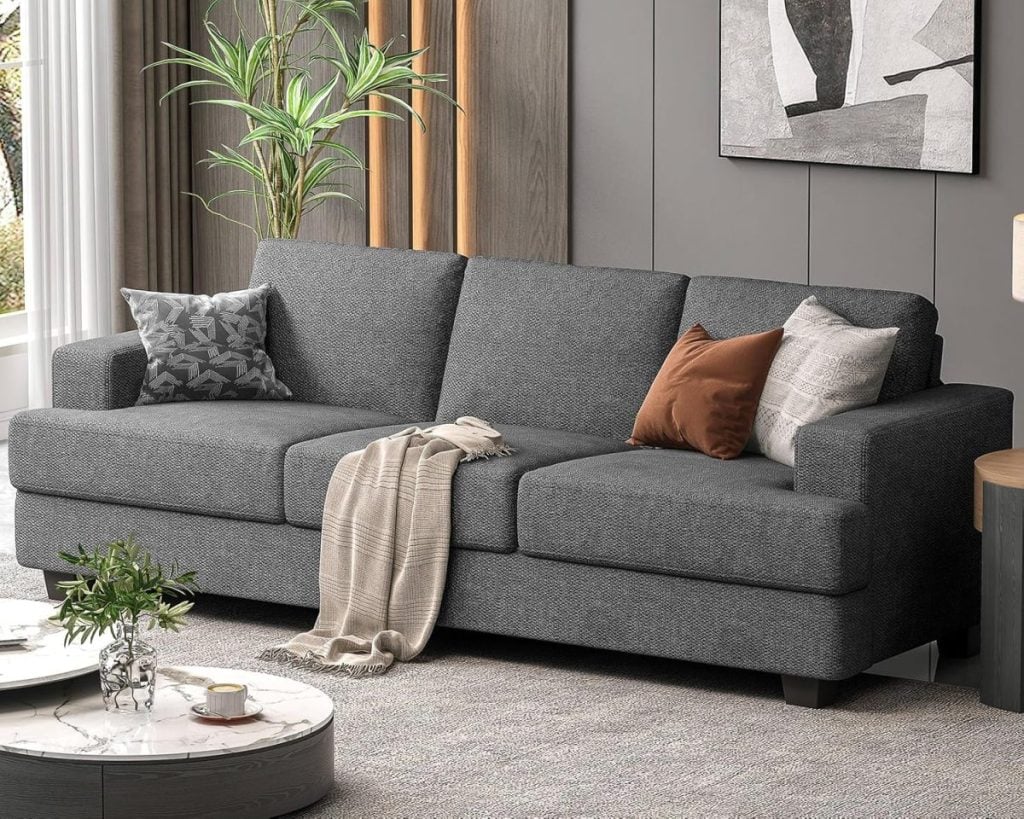 A gray sofa with multiple pillows and a draped blanket is set in a modern living room with a potted plant, marble coffee table, and abstract art on the wall.