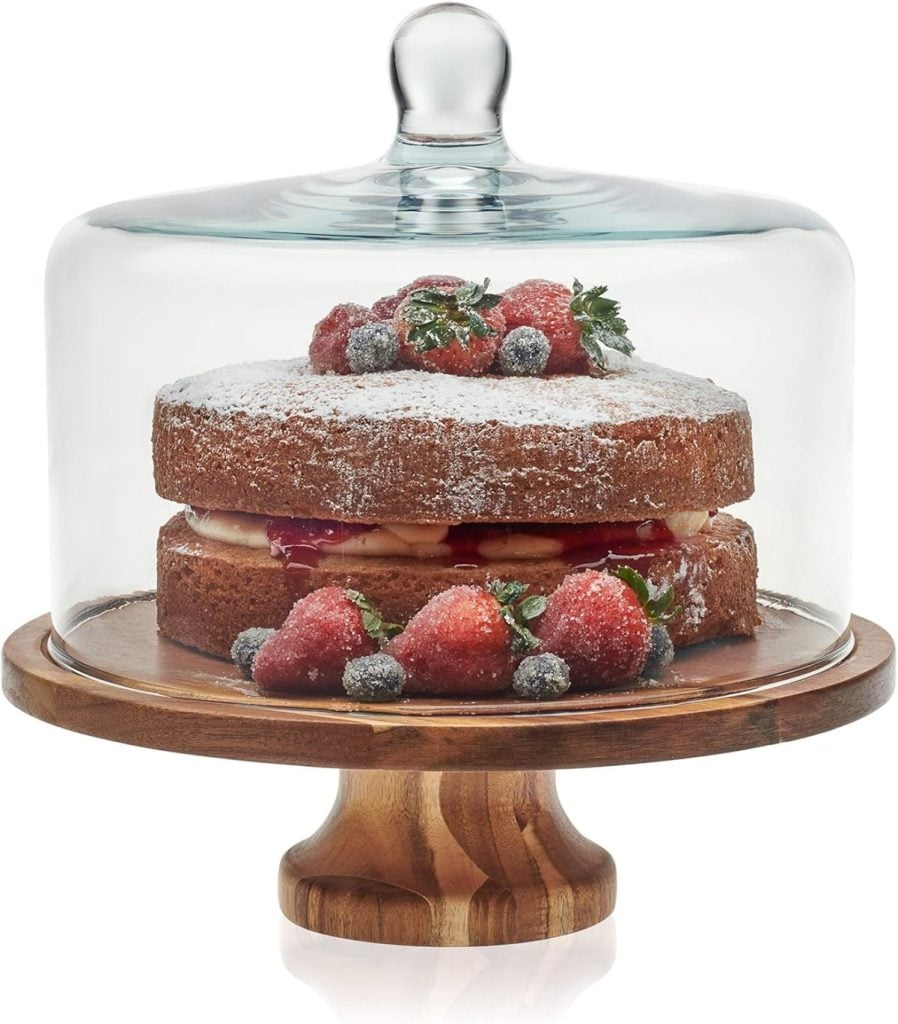 A layered cake with powdered sugar and mixed berries sits on a wooden cake stand, covered by a glass dome, offering an irresistible treat that rivals having pie for breakfast.