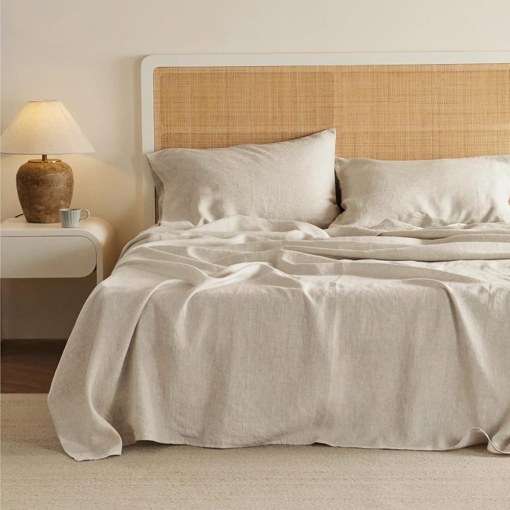 A neatly made bed with beige linen sheets and pillows, beside a white nightstand with a ceramic lamp and a cup.