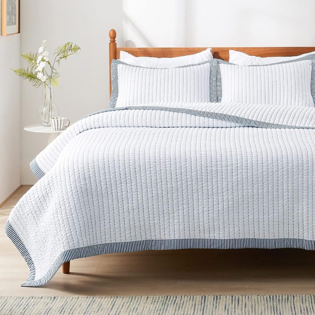 A neatly made bed with a white, striped quilt and matching pillows, a wooden headboard, a small side table with a vase holding greenery and white flowers, and a patterned rug on the floor.