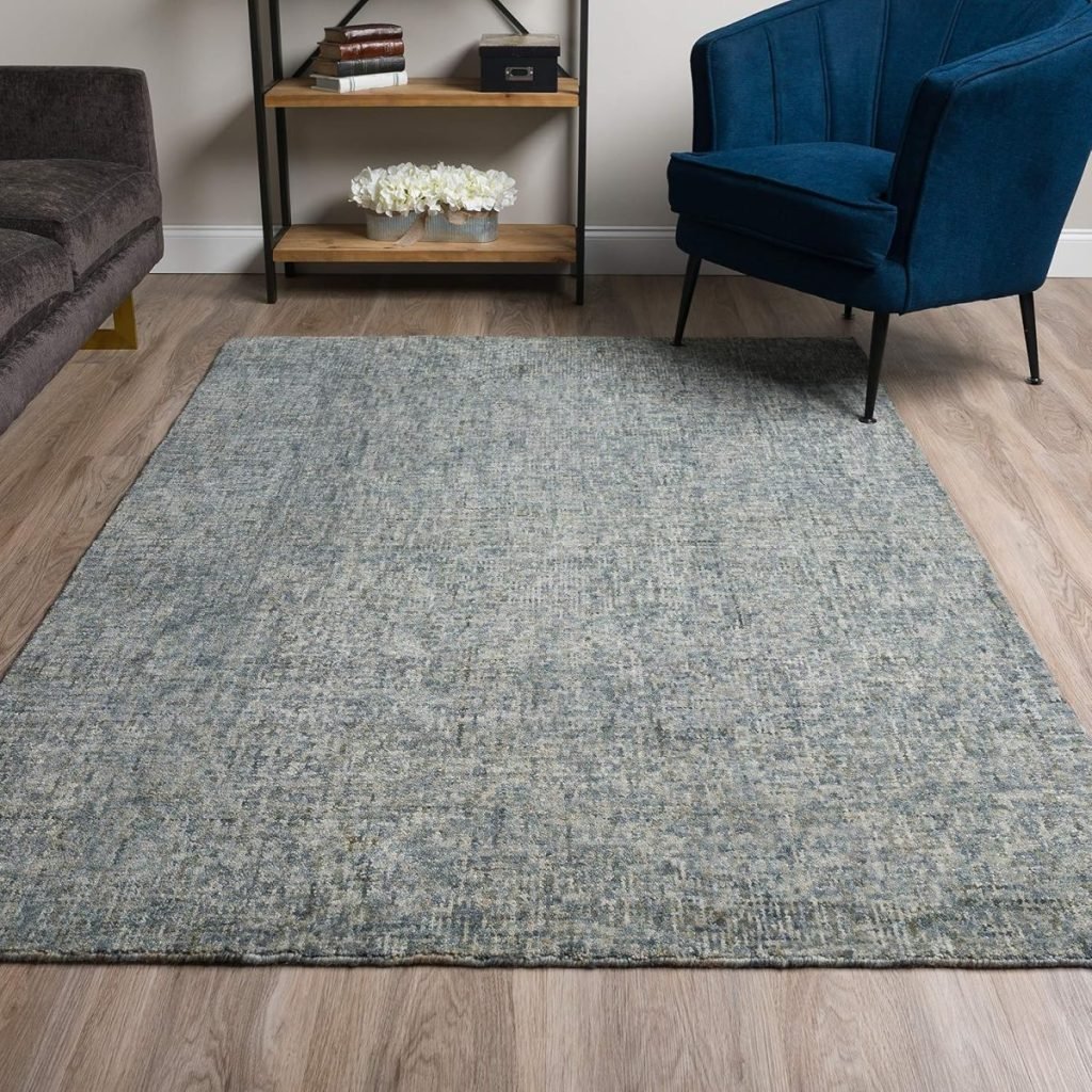 A gray textured rug on a light wood floor between a gray couch and a blue armchair, with a black metal and wood shelf holding books and flowers in the background.