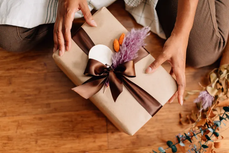 Person holding a gift wrapped in brown paper with a dark ribbon and decorative dried flowers on a wooden floor.