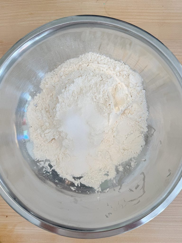 A stainless steel bowl containing a dry mixture of flour and sugar on a wooden surface.