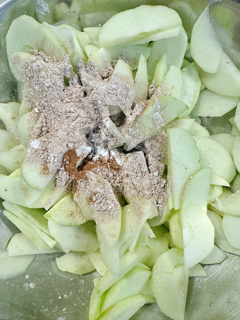 Sliced green apples topped with a mixture of flour, sugar, and spices in a metal bowl.