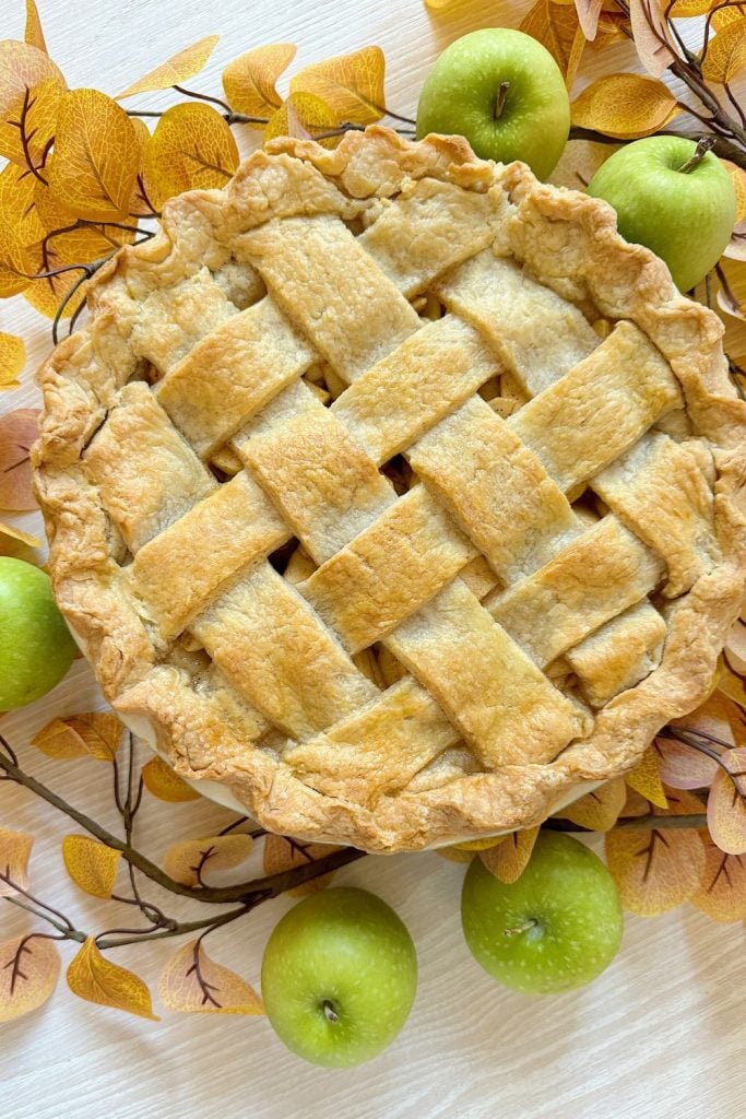 A lattice-top pie surrounded by green apples and autumn leaves on a light wooden surface.