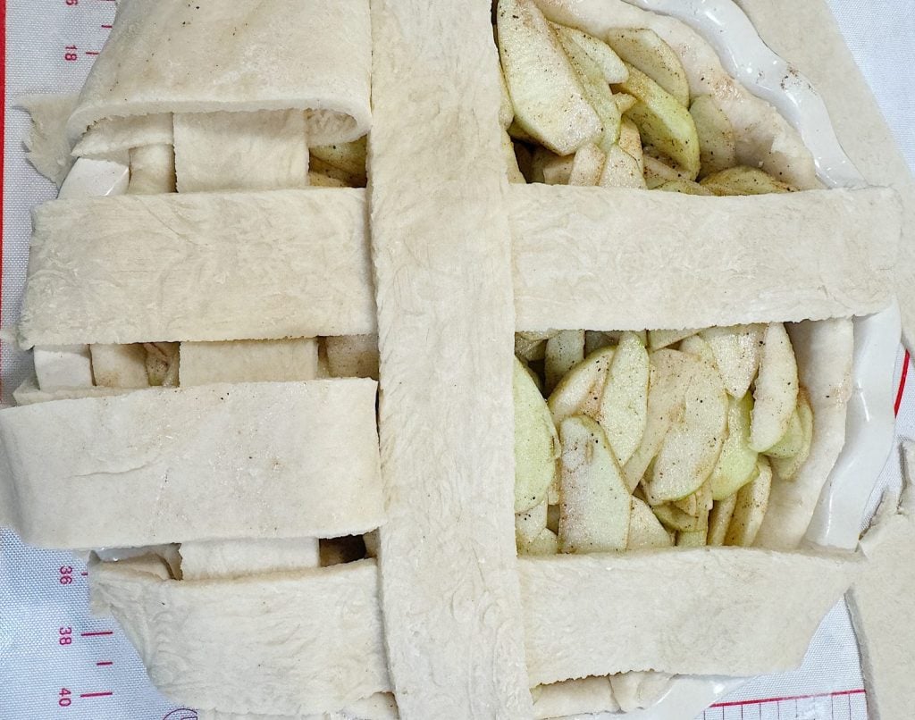 Unbaked lattice apple pie with partially arranged dough strips over apple filling on a pastry mat.