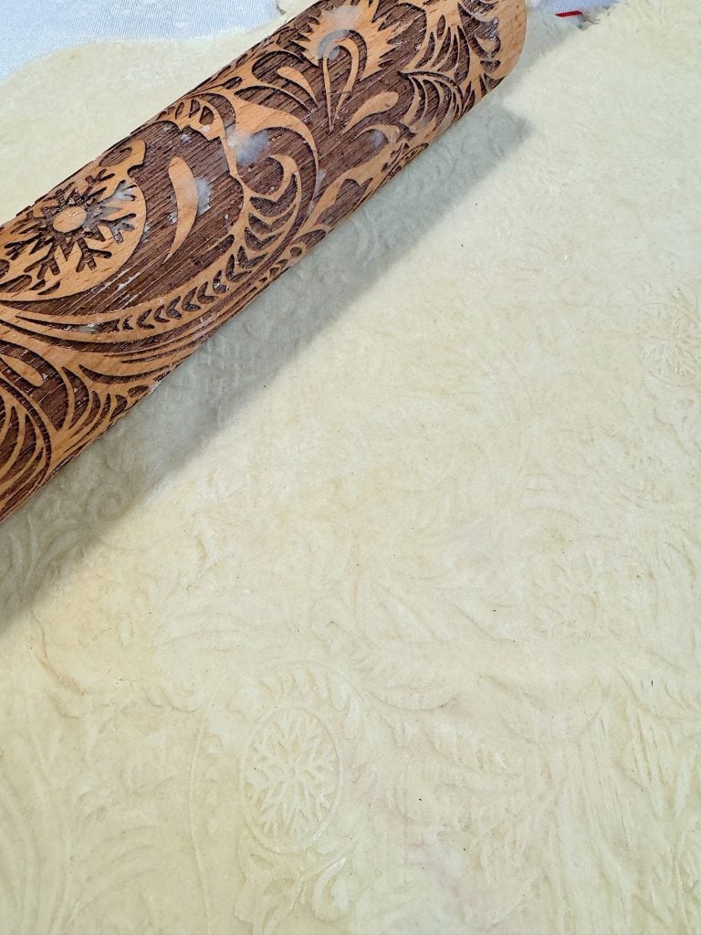 Embossed wooden rolling pin on textured dough, featuring floral and leaf patterns.