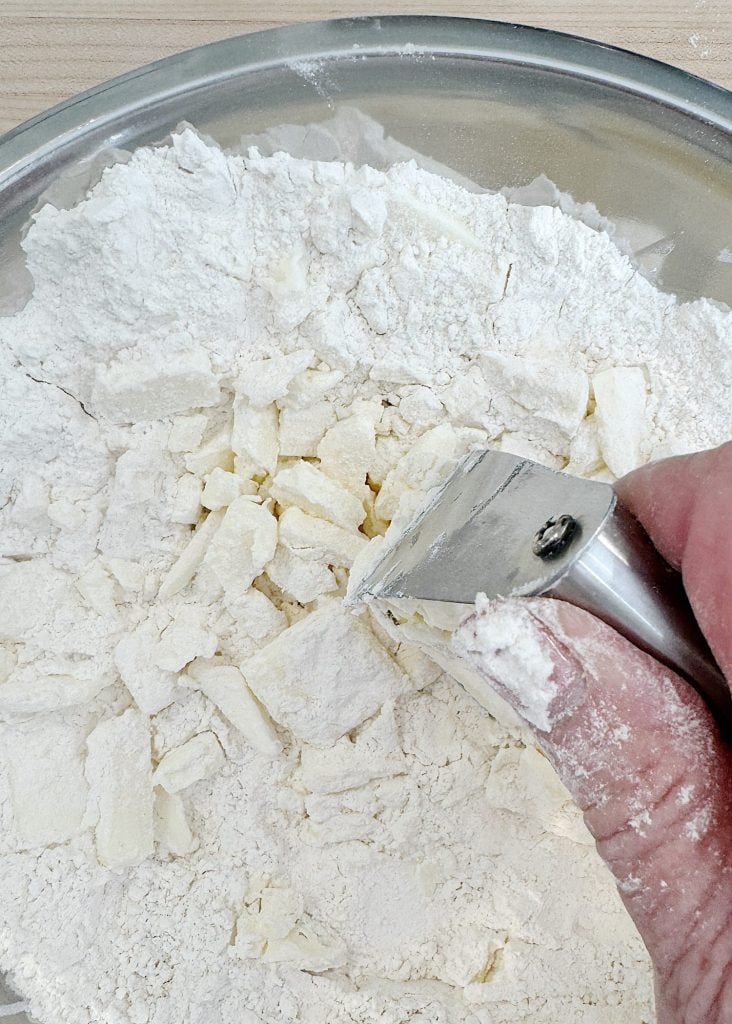 A hand using a pastry cutter to mix butter into flour in a metal bowl.