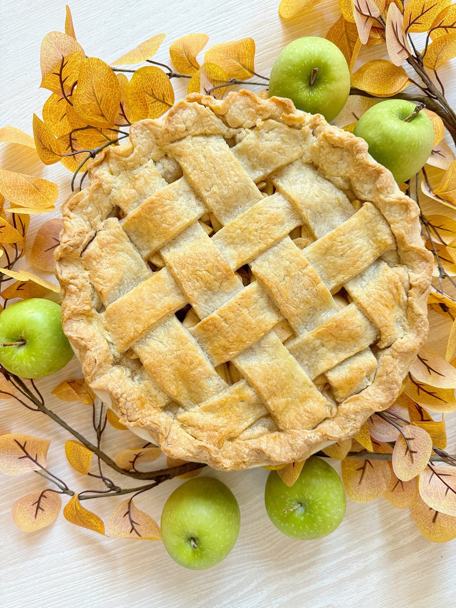 A lattice-top pie surrounded by green apples and autumn leaves on a light-colored surface.