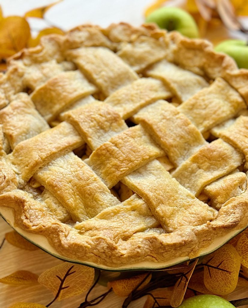 A freshly baked lattice-topped apple pie on a wooden table, surrounded by autumn leaves and green apples.