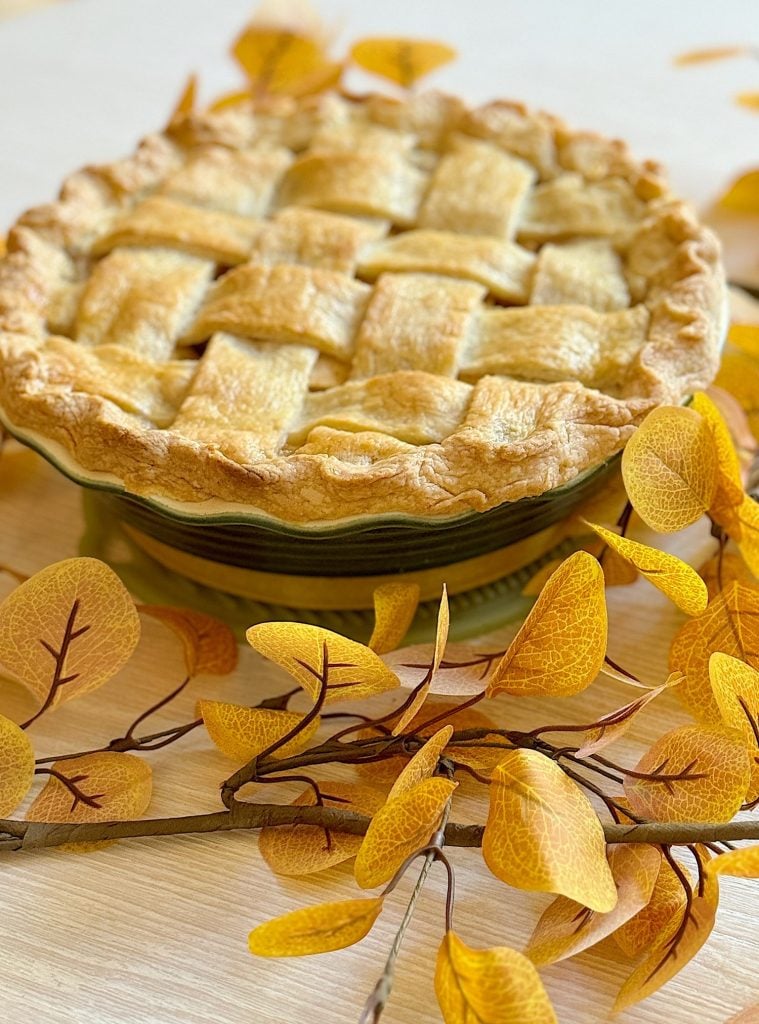 A lattice-topped pie sits on a table surrounded by yellow autumn leaves.