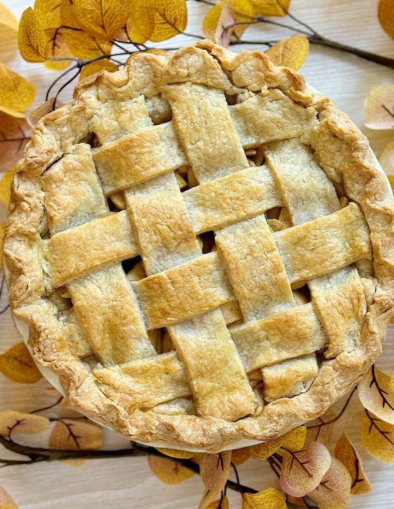 A lattice-topped pie surrounded by autumn leaves on a light wooden surface.