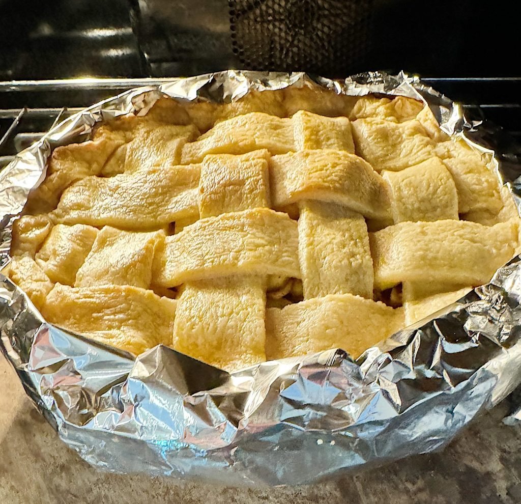 A homemade pie with a lattice crust wrapped in aluminum foil is baking in an oven.