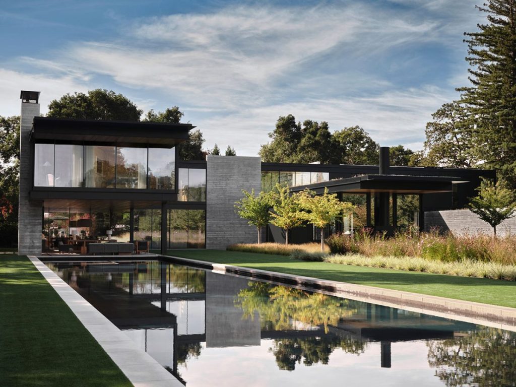 Modern house with large glass windows surrounded by trees, featuring a long rectangular pool reflecting the architecture and sky.