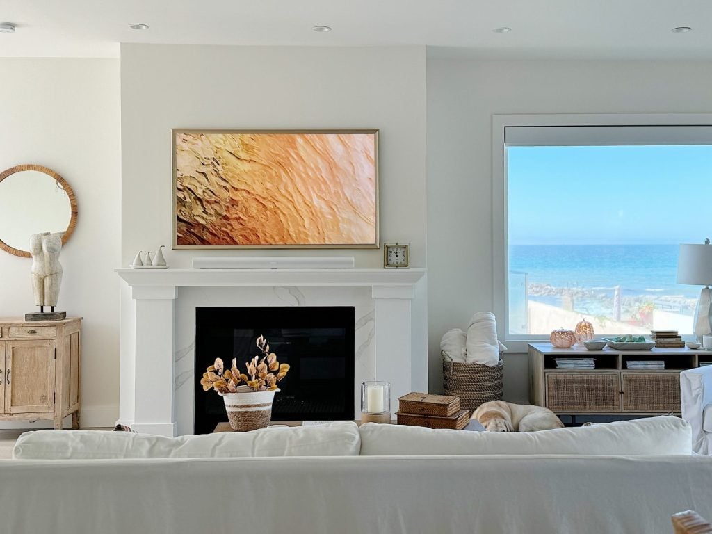 A living room with a white sofa, fireplace, and framed art. Large window reveals an ocean view. Natural decor elements like a plant and woven baskets are present.