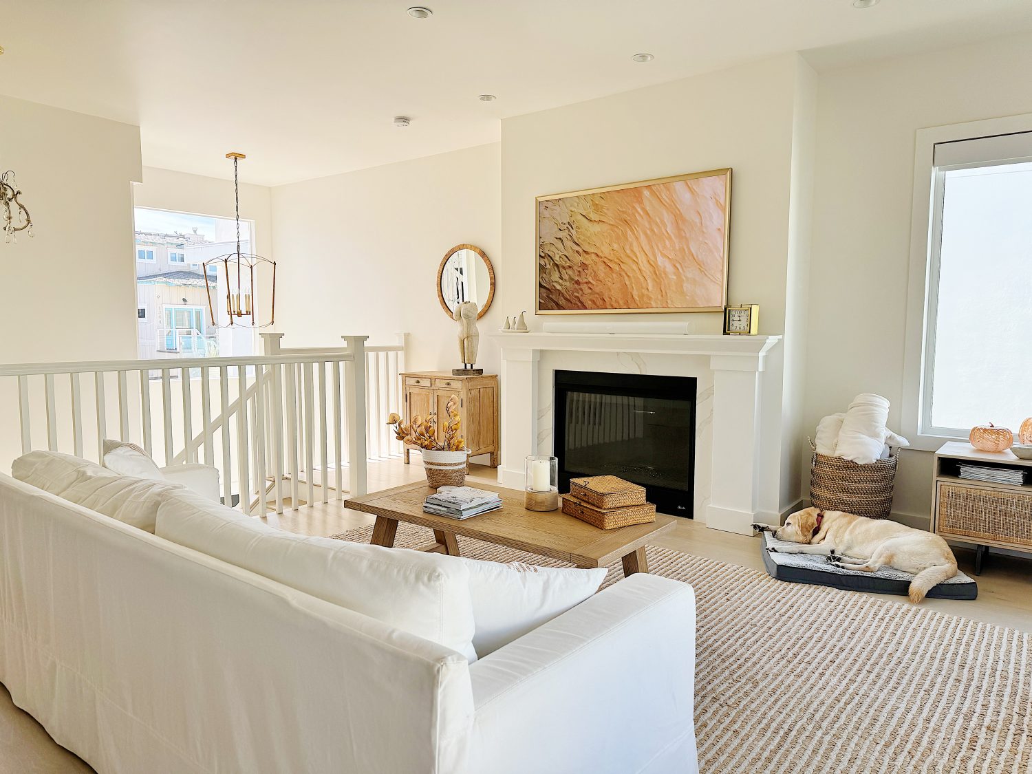 Living room with a white sofa, coffee table, dog in a bed, fireplace, wall art, and a window. Neutral decor with a round mirror above a sideboard.