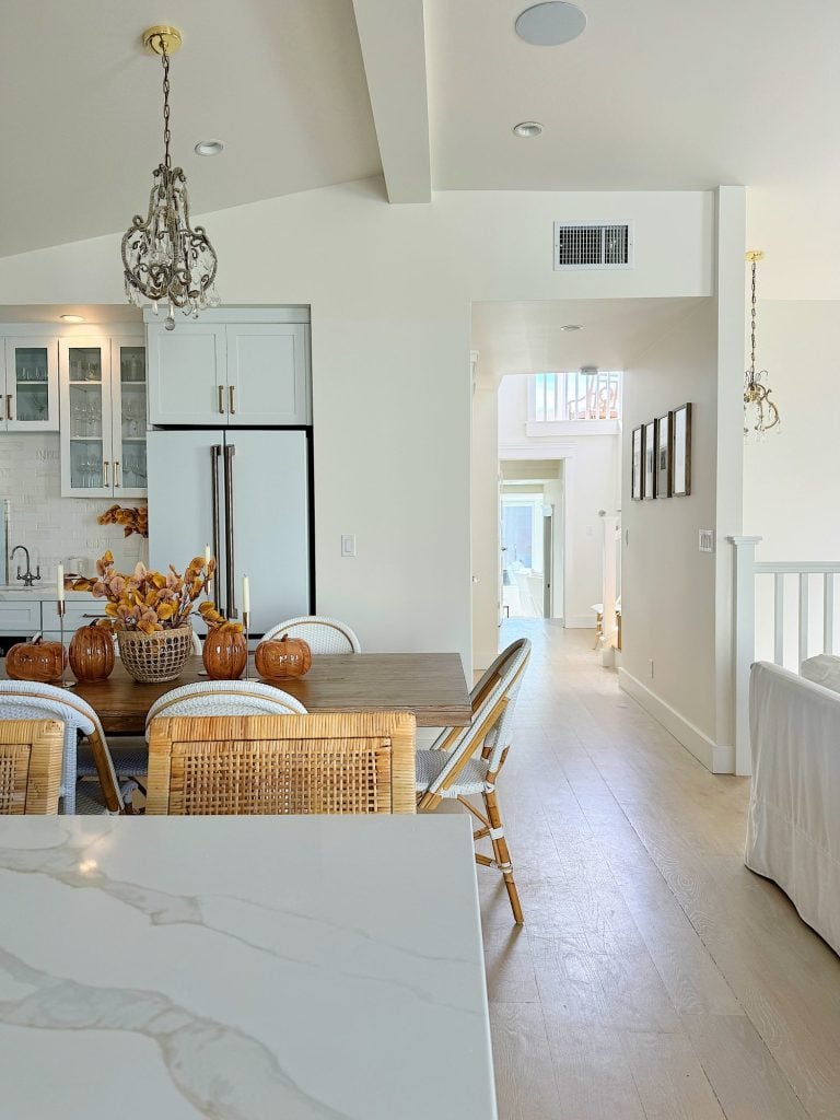 A modern kitchen and dining area featuring a wooden table, wicker chairs, and a white countertop with a decorative chandelier overhead. In the background, a hallway leads to a well-lit room.