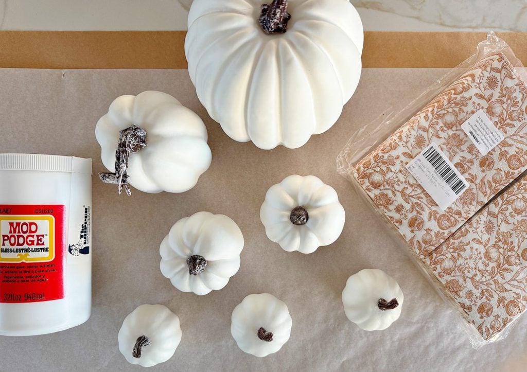 Various sized white decorative pumpkins, a container of Mod Podge, and a pack of patterned paper napkins are arranged on a surface.