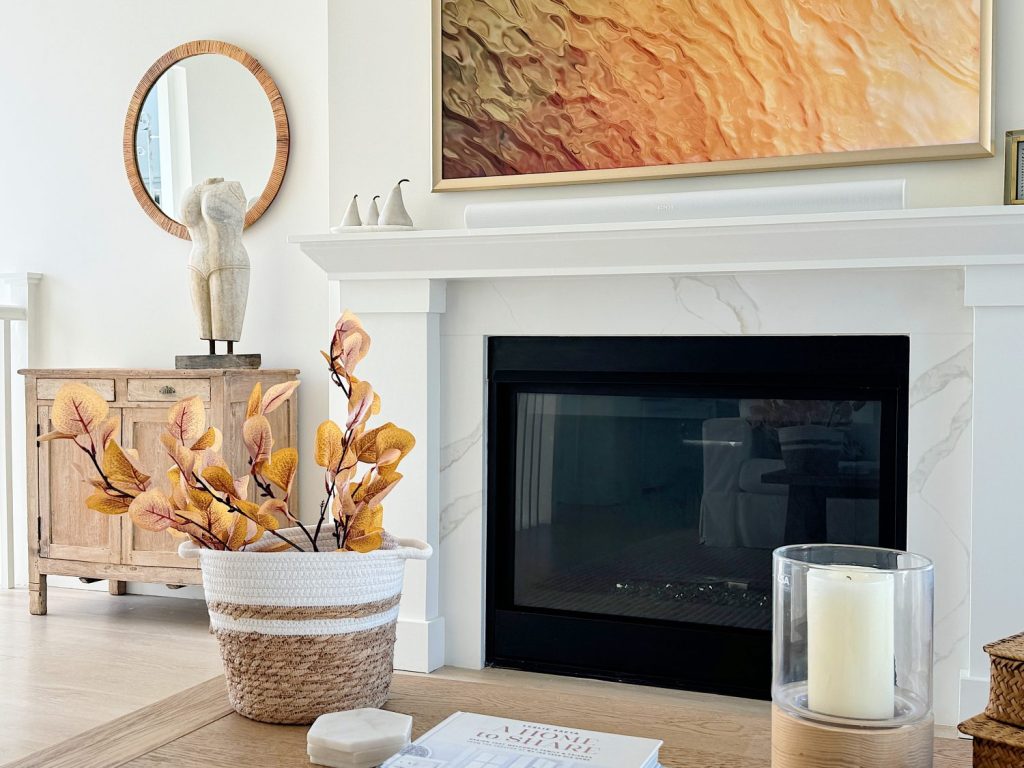 A cozy living room with a fireplace, a round mirror, a wooden cabinet, and a basket of autumn leaves on the coffee table.