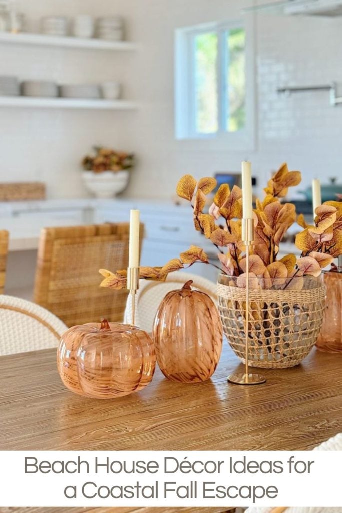 A dining table with autumn-themed decor, including glass pumpkin ornaments, a woven vase with orange leaves, and two candlesticks. White chairs and shelves are in the background.