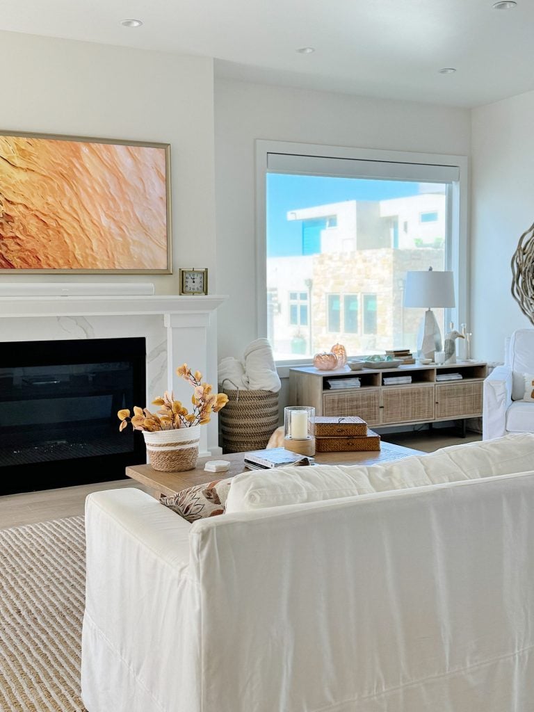 A modern living room with a white sofa, fireplace, abstract art, and large window. Natural light fills the space, highlighting neutral tones and a minimalist design.