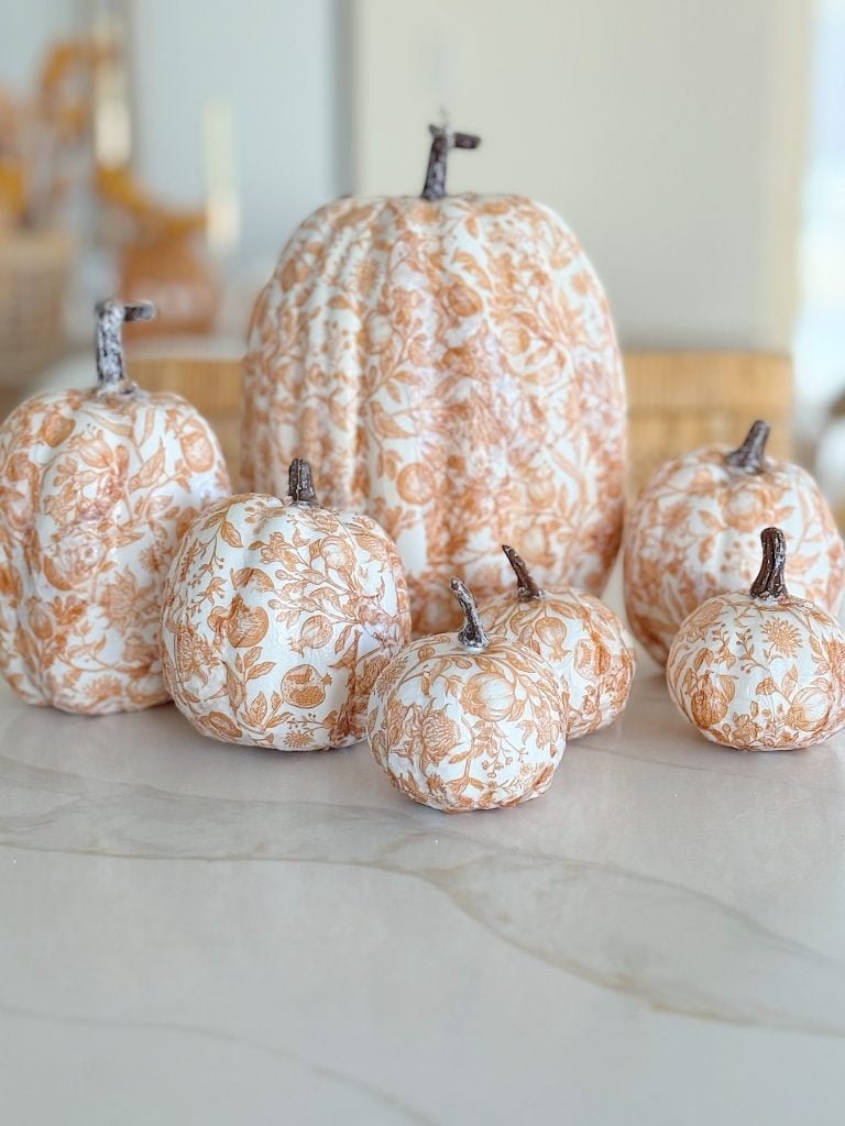 A group of pumpkins decorated with an intricate floral pattern in beige and white are displayed on a smooth, light-colored surface.