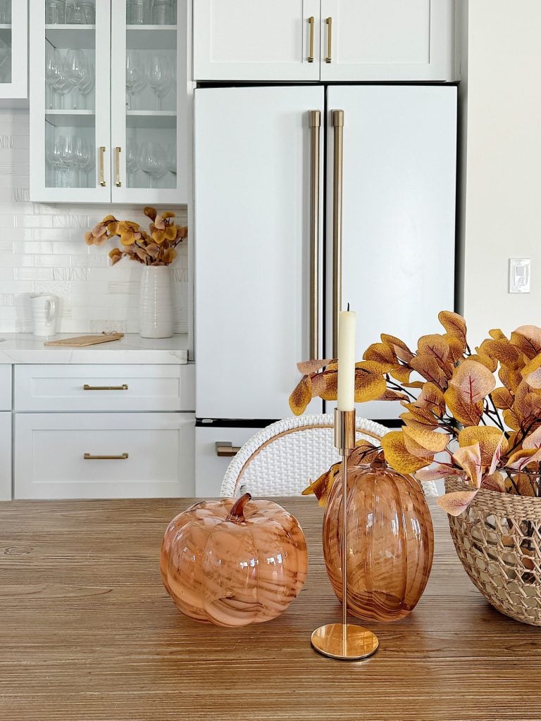 A kitchen with white cabinets and a refrigerator, featuring a wooden table decorated with orange glass pumpkins, a candle holder, and a basket of autumn leaves.