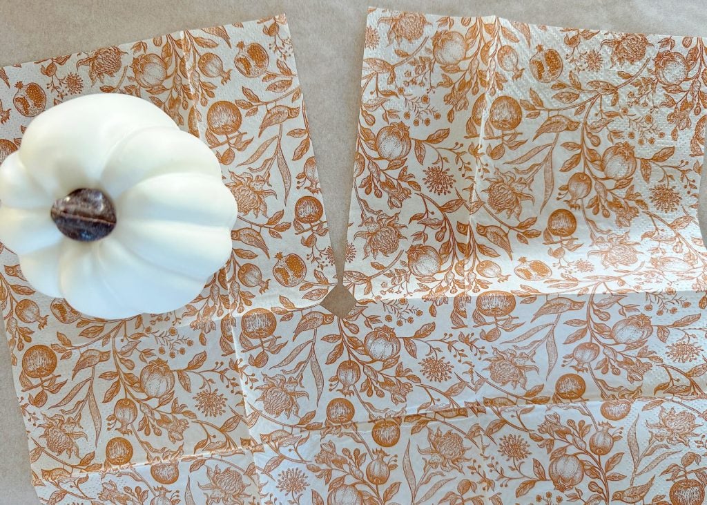 A small white pumpkin sits on decorative paper with orange floral and botanical patterns on a beige surface.