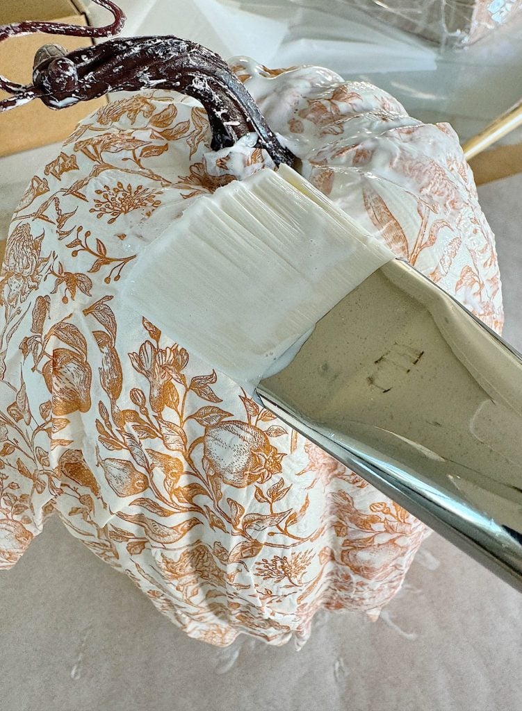 A paintbrush applies white paint over a decorative pumpkin with a floral pattern.