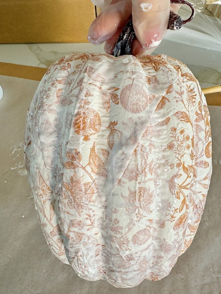 A hand holds a decorative pumpkin covered in white paint with orange floral patterns on a beige background.