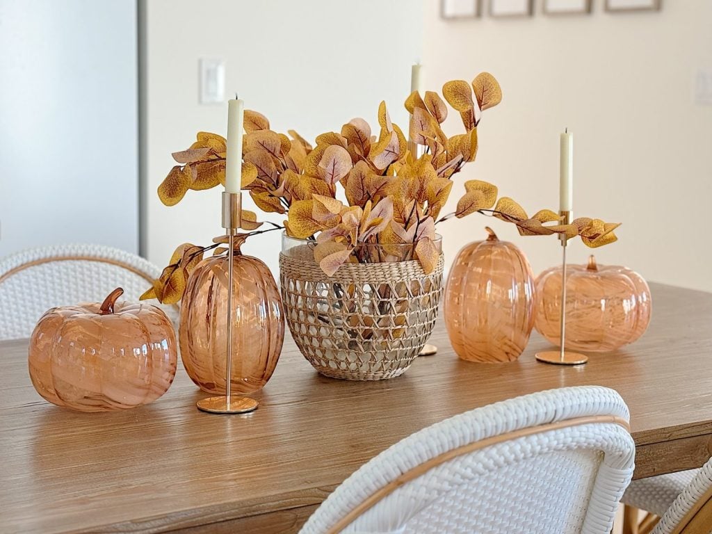 Table with autumn-themed decor, featuring orange glass pumpkins, a woven basket with yellow leaves, and two candle holders with white candles. White woven chairs surround the table.