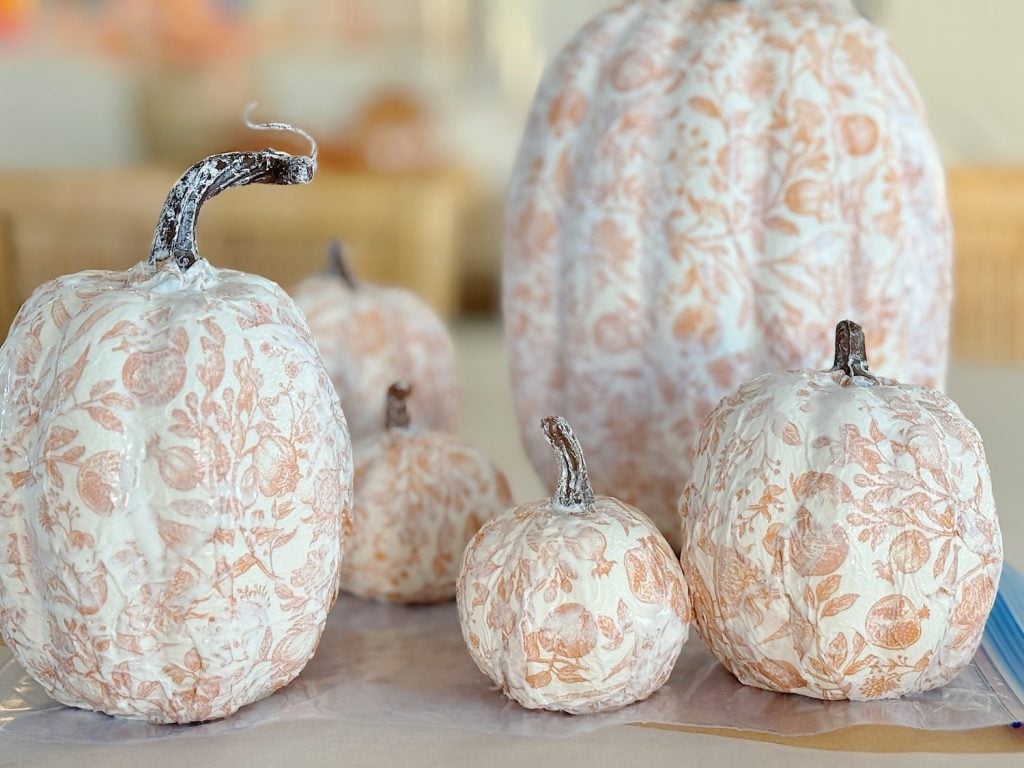 Five pumpkins are decorated with white and beige floral patterns. They are arranged on a flat surface.