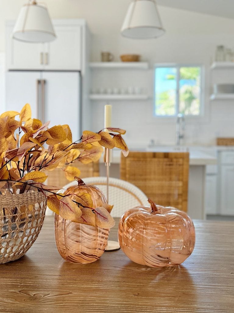 A kitchen table features decorative glass pumpkins and a basket with autumn leaves. The background shows a bright, modern kitchen with white cabinets and wicker accents.