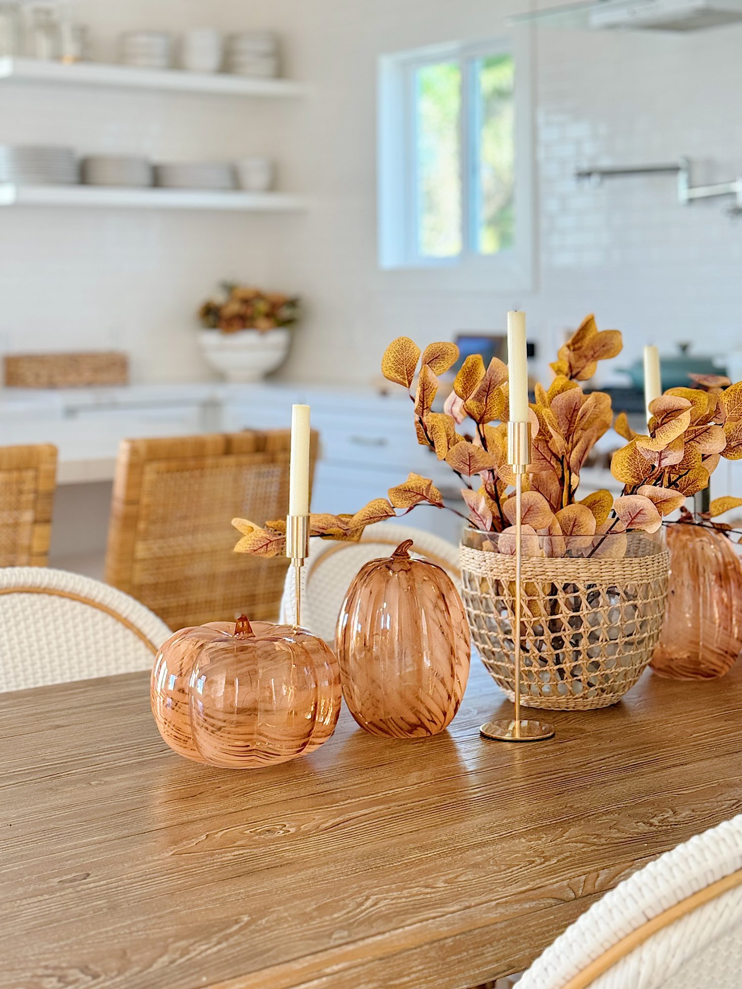 A wooden table with two glass pumpkins, a basket with yellow leaves, and a gold candlestick in a bright kitchen setting.