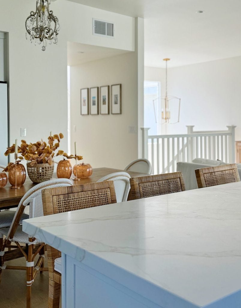 A modern kitchen and dining area with white and wicker furniture, a marble countertop, decorative pumpkins, and framed pictures on the wall. A chandelier and pendant light are visible.