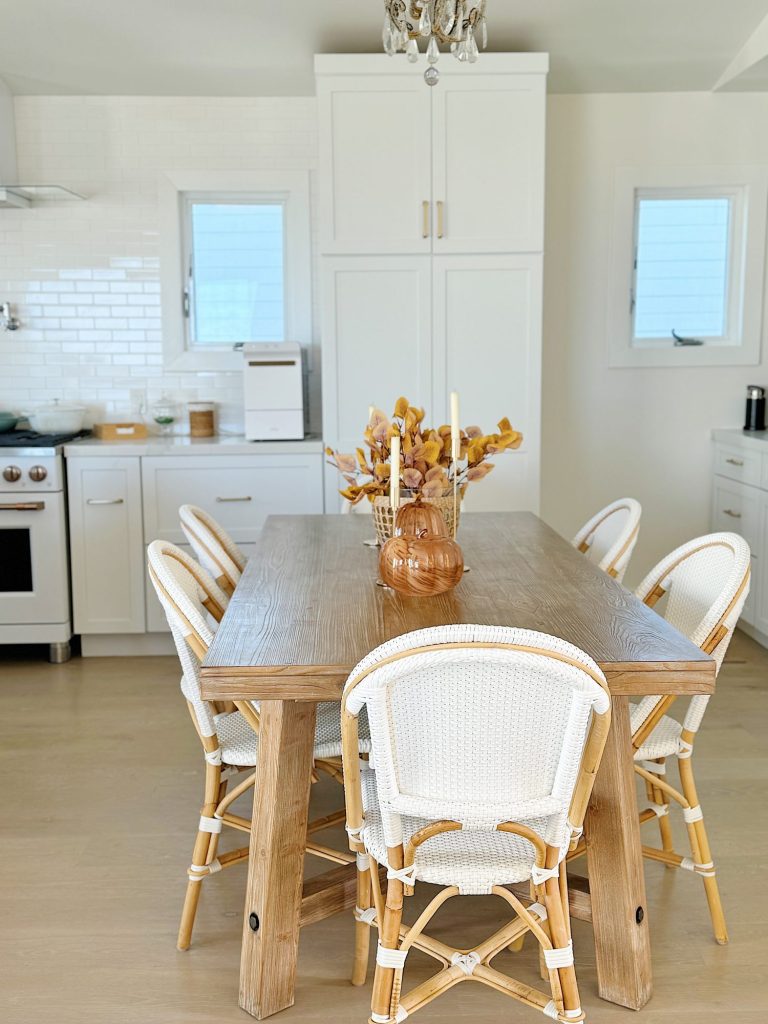 Dining room with a wooden table, six white wicker chairs, a centerpiece with candles and dried plants, white cabinets, and a kitchen area with a stove and oven in the background.