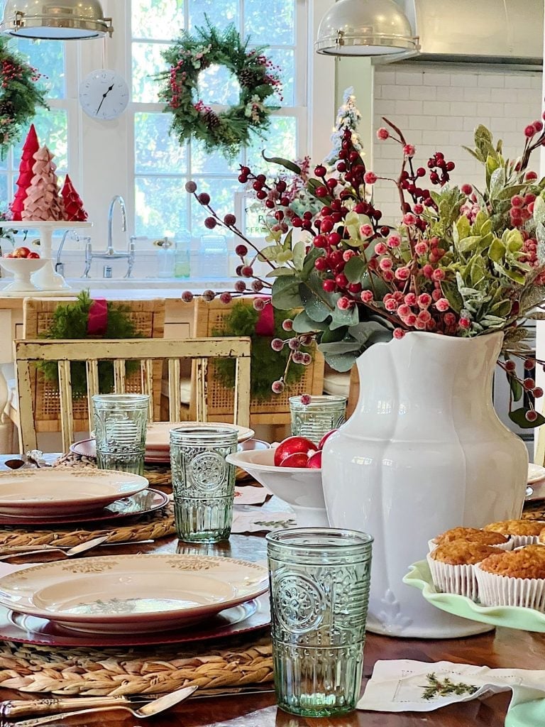 A festive dining table is set with green glassware, plates, and a large white vase with red berry branches. Christmas wreaths decorate the windows in the background.