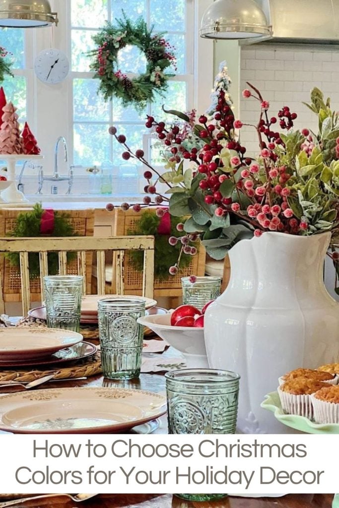 Festive dining table with green glassware, a white vase of red and green foliage, and holiday decorations. A window with a wreath in the background.