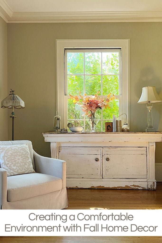 Living room with a white armchair, a vintage white console table, and a window view. Decor includes a lamp, flower arrangement, and small ornaments. Text reads: "Creating a Comfortable Environment with Fall Home Decor.