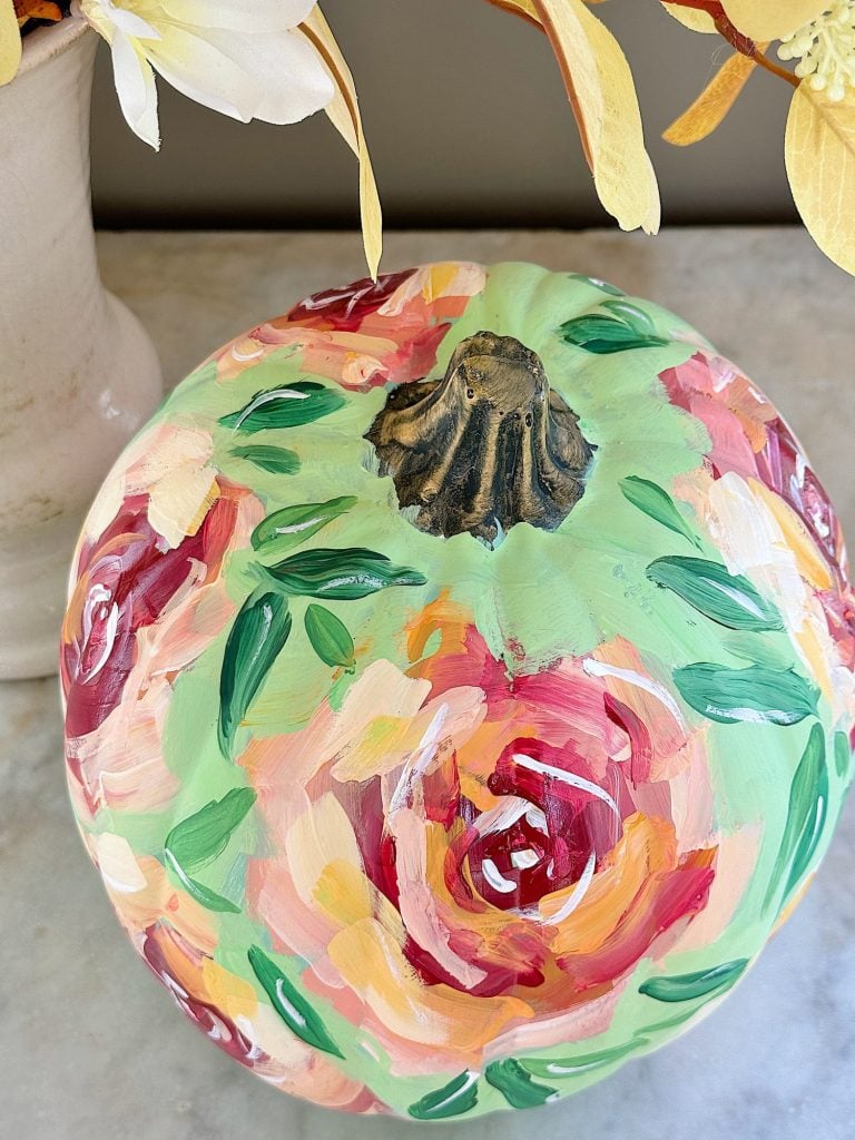 A pumpkin painted with colorful, abstract floral patterns sits on a marble surface next to a vase of flowers.