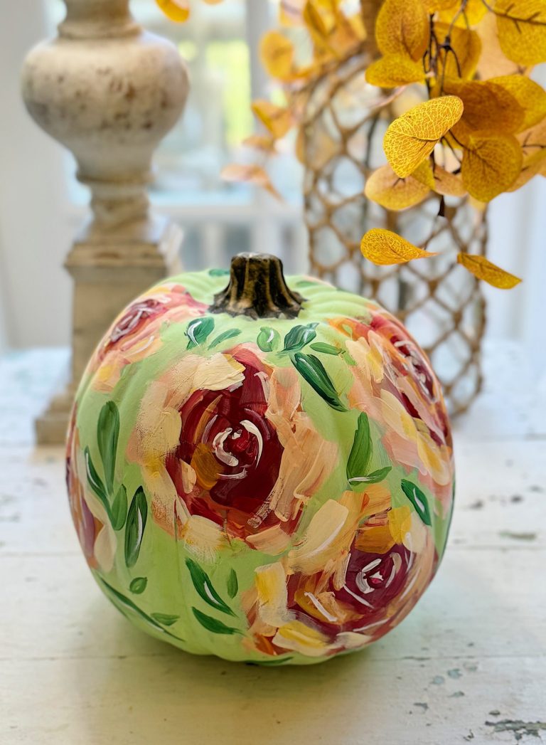 Decorative pumpkin painted with colorful floral designs in front of a vase with yellow leaves.