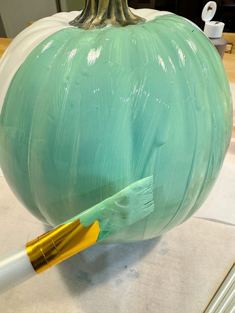 A large pumpkin being painted light teal with a paintbrush.