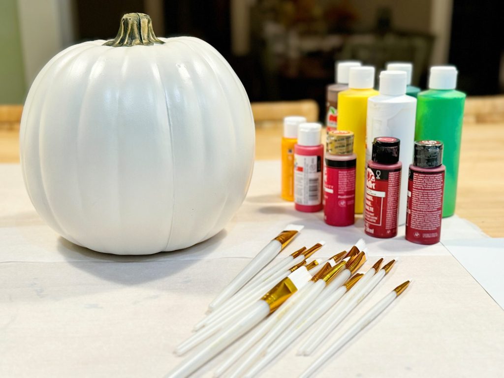 White pumpkin with multiple paint bottles and brushes on a table prepared for painting.