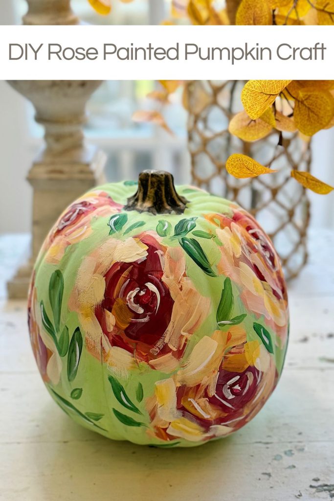 A pumpkin painted with rose designs in pink, red, and green, placed on a table with autumn leaves nearby.